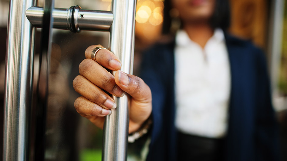 En hand som tar tag i ett dörrhandtag, i bakgrunden en kontorsmiljö och en person i arbetskläder.